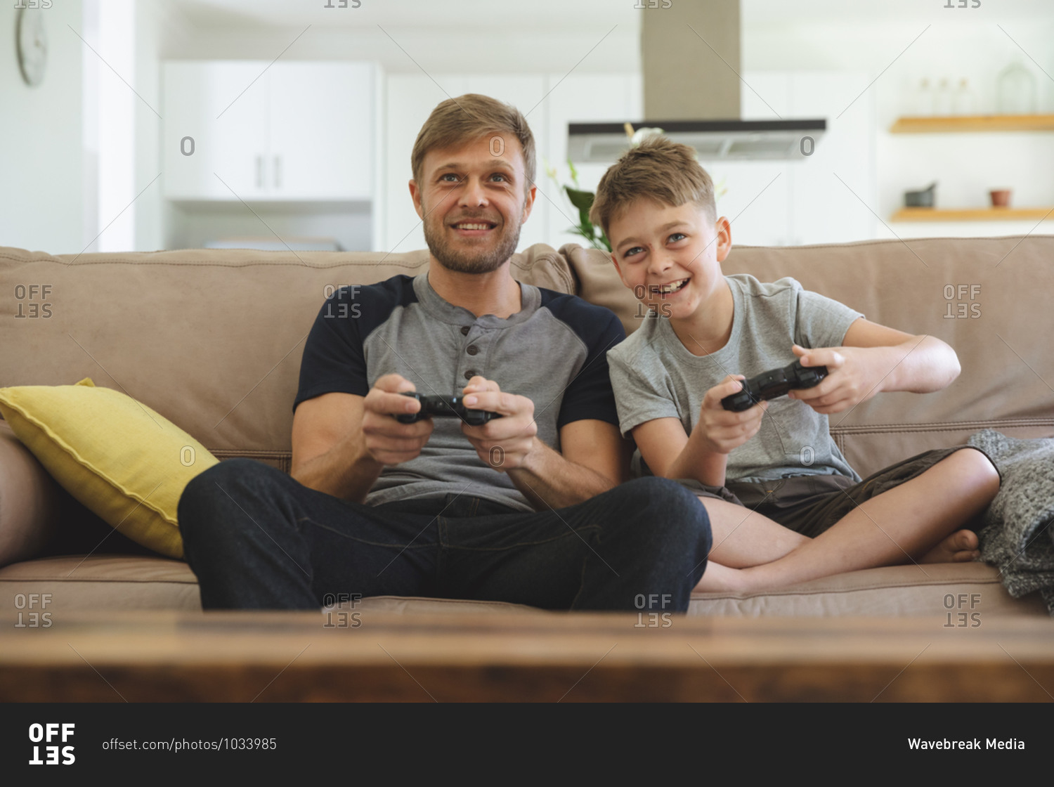 Caucasian man at home with his son together, sitting on sofa in living room, playing video games, smiling. Social distancing during Covid 19 Coronavirus quarantine lockdown.