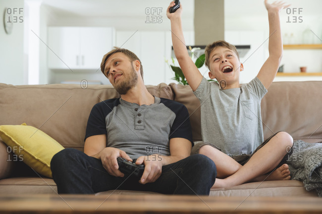 Caucasian man at home with his son together, sitting on sofa in living room, playing video games, boy cheering victory with arms up. Social distancing during Covid 19 Coronavirus quarantine lockdown.