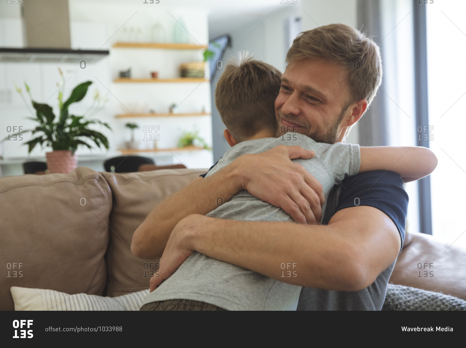 Caucasian man at home with his son together, sitting on sofa in living room, embracing each other, smiling. Social distancing during Covid 19 Coronavirus quarantine lockdown.