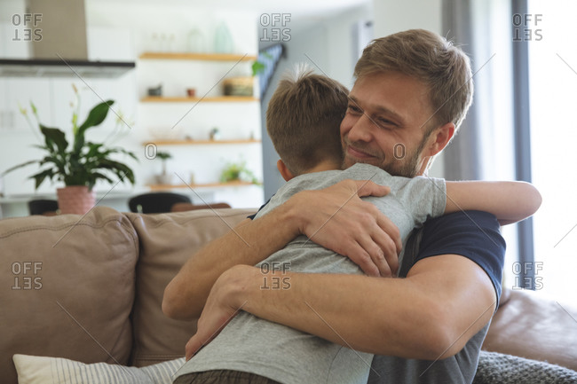 Caucasian man at home with his son together, sitting on sofa in living room, embracing each other, smiling. Social distancing during Covid 19 Coronavirus quarantine lockdown.