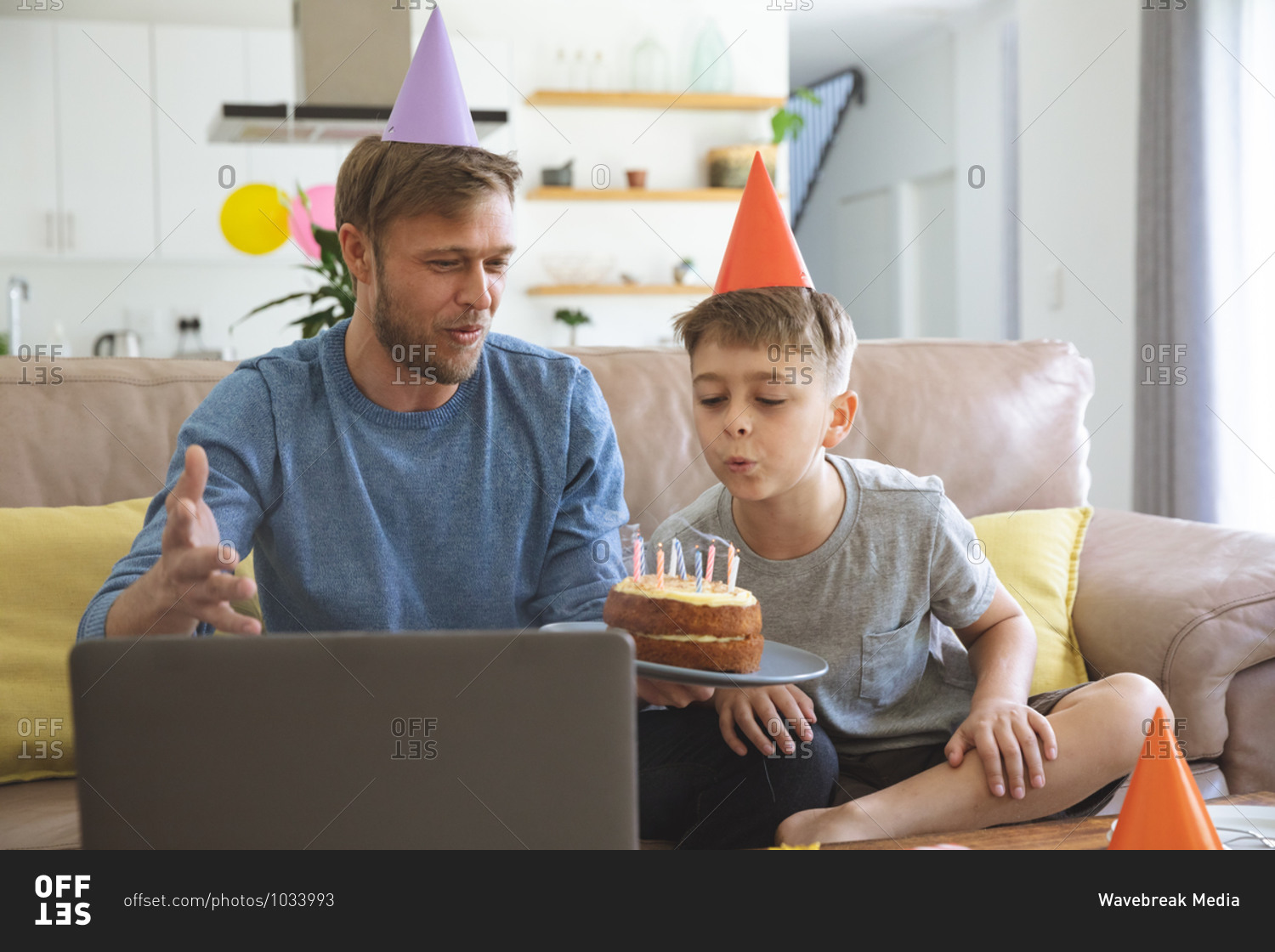 Caucasian man spending time at home with his son together, blowing candles on  birthday cake using laptop computer for video chat. Social distancing during Covid 19 Coronavirus quarantine lockdown.
