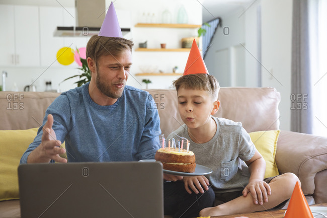 Caucasian man spending time at home with his son together, blowing candles on  birthday cake using laptop computer for video chat. Social distancing during Covid 19 Coronavirus quarantine lockdown.