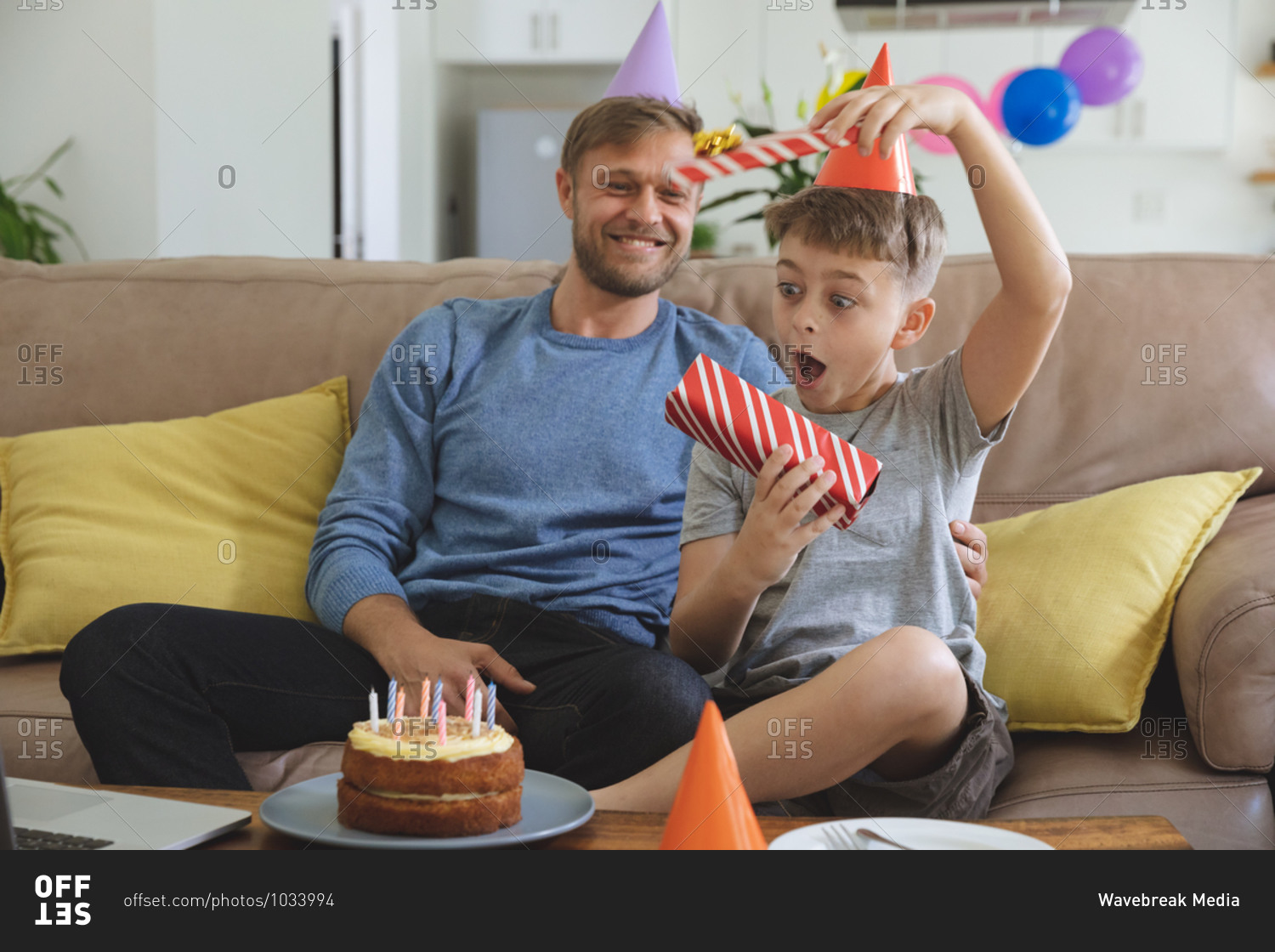 Caucasian man spending time at home with his son together, wearing party hats boy receiving birthday present. Social distancing during Covid 19 Coronavirus quarantine lockdown.