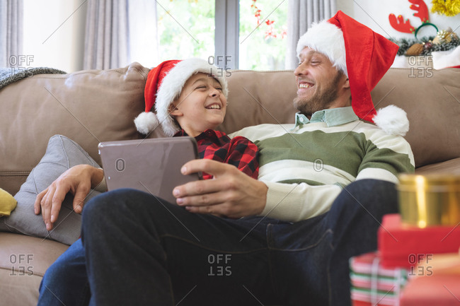 Caucasian man at home with his son at Christmas, wearing Santa hats sitting in living room using digital tablet. Social distancing during Covid 19 Coronavirus quarantine lockdown.