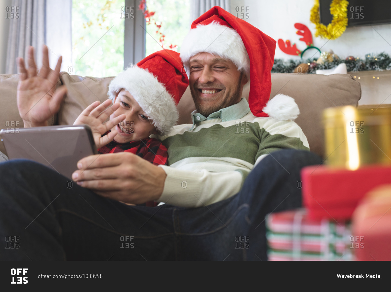 Caucasian man at home with his son at Christmas, wearing Santa hats sitting on sofa in living room, using digital tablet, waving. Social distancing during Covid 19 Coronavirus quarantine lockdown.