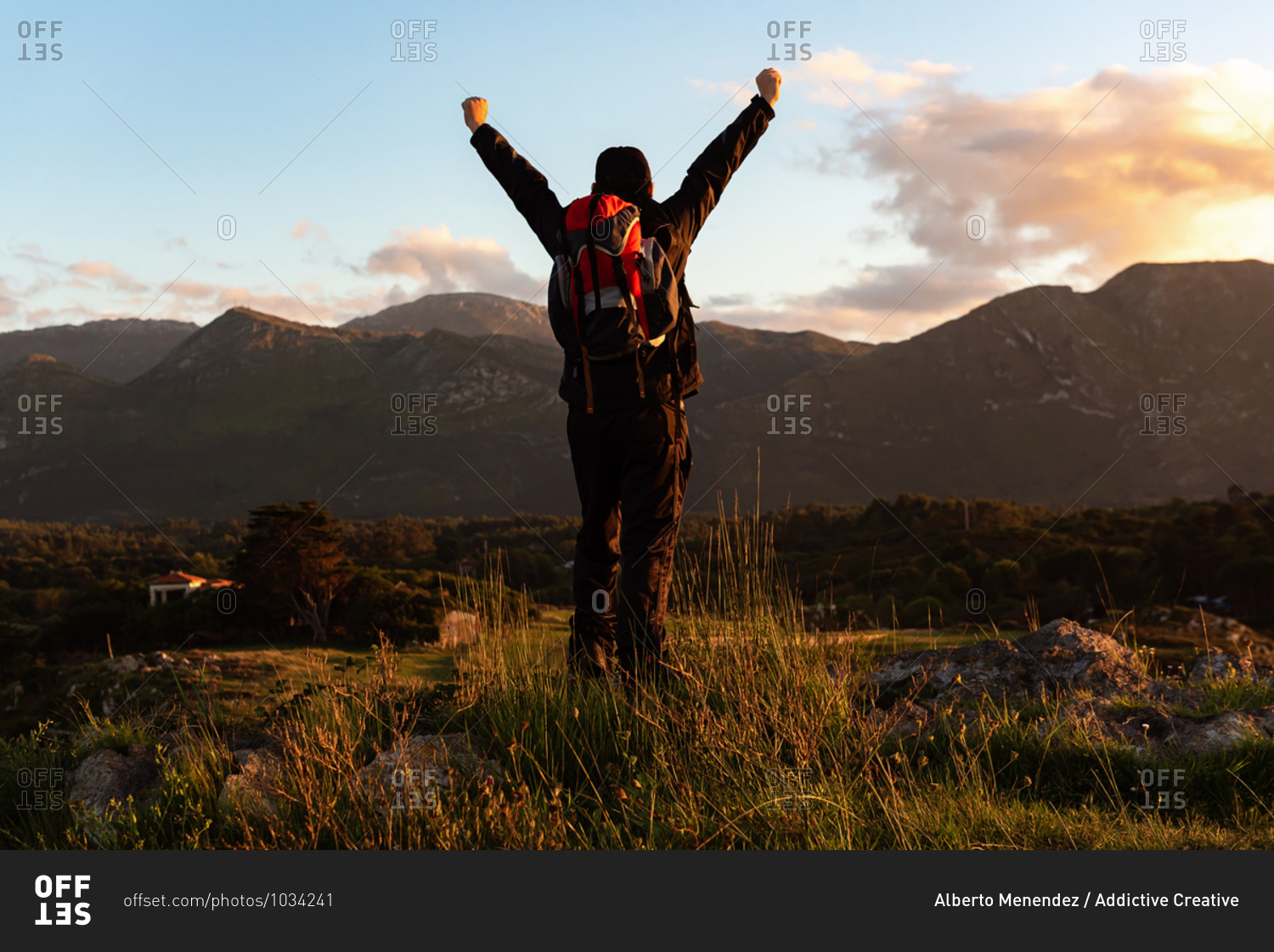Back view of unrecognizable male traveler with backpack standing on hill and admiring scenery of mountain ridge under sunset sky