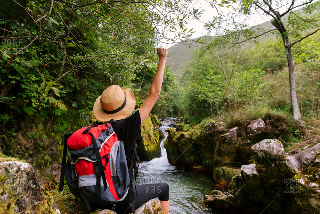 Back view of unrecognizable hiker with backpack celebrating goal achievement with fist up while standing near mountain river in woods