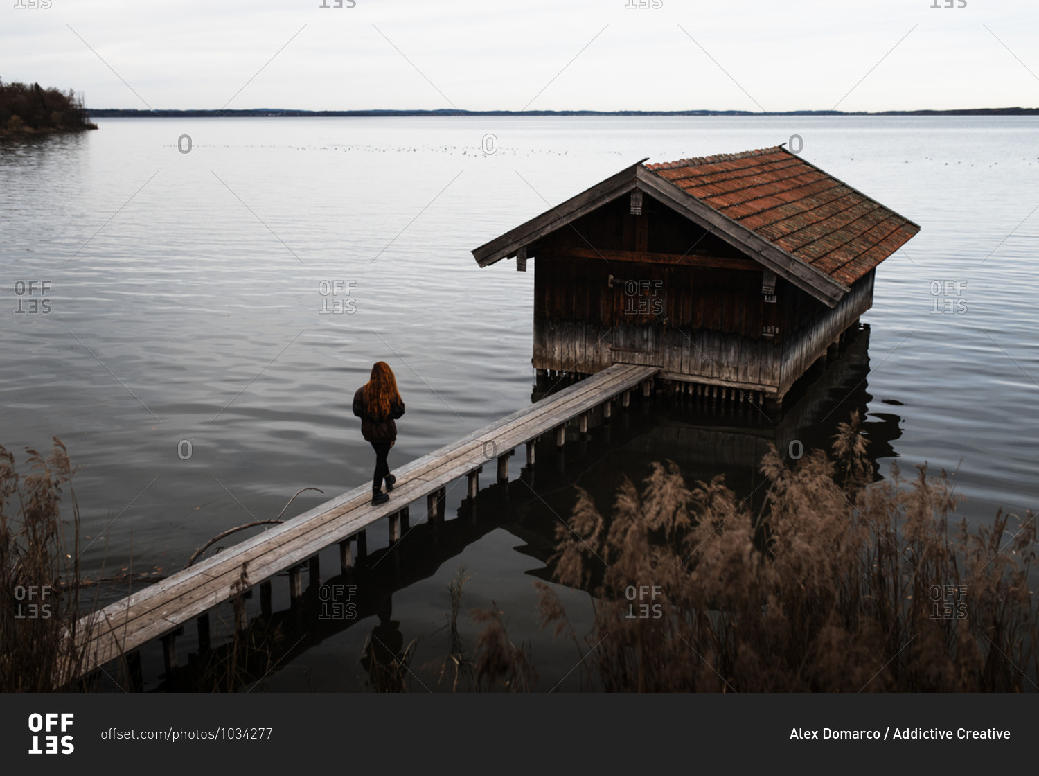 Unrecognizable female traveler in warm clothes standing on wooden plank pier near shabby shed located at lake in gloomy autumn day