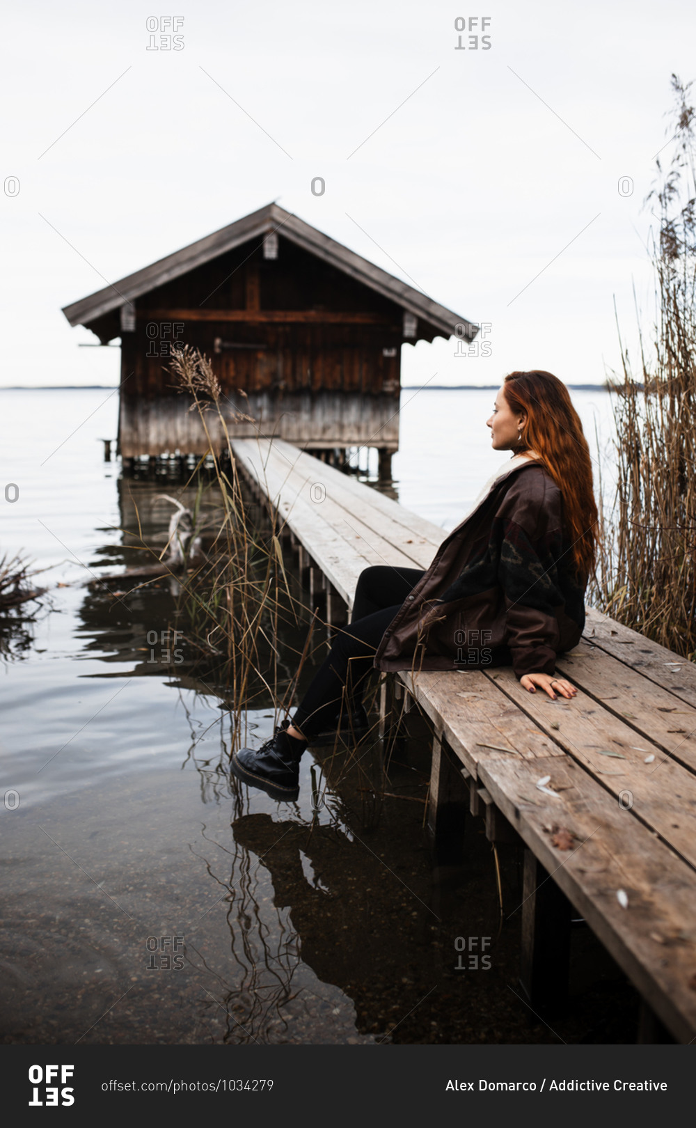 Female traveler in warm clothes sitting on wooden plank pier near shabby shed located at lake in gloomy autumn day