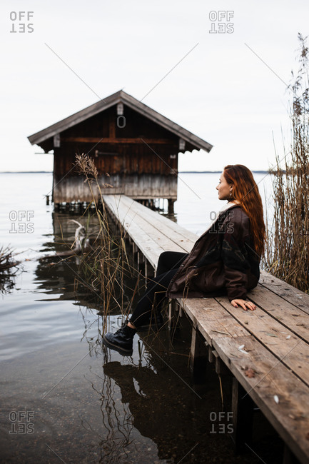 Female traveler in warm clothes sitting on wooden plank pier near shabby shed located at lake in gloomy autumn day