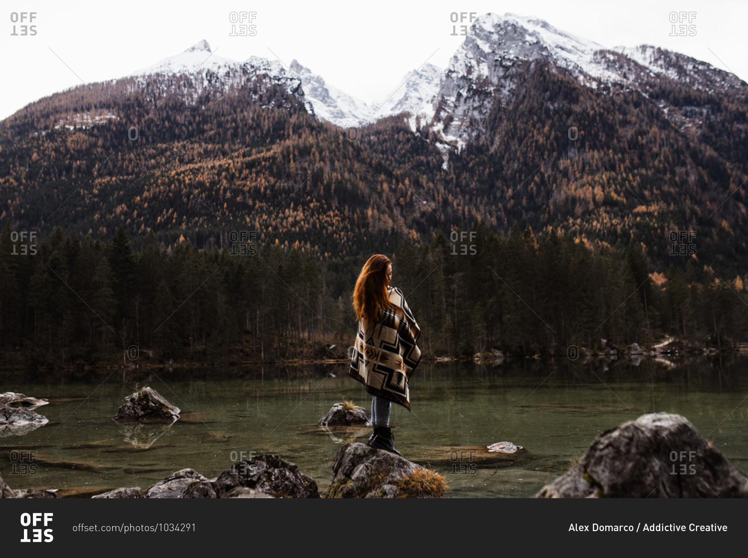Side view of unrecognizable distant female traveler in warm outfit standing on rocky shore of lake and observing amazing scenery of autumn forest and snowy mountains in cloudy day