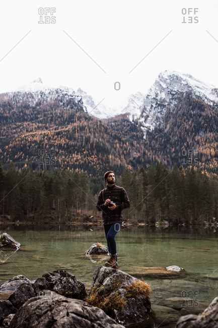 Young male photographer in warm jacket standing on stone of lake against blurred mountains and taking pictures with photo camera in autumn day
