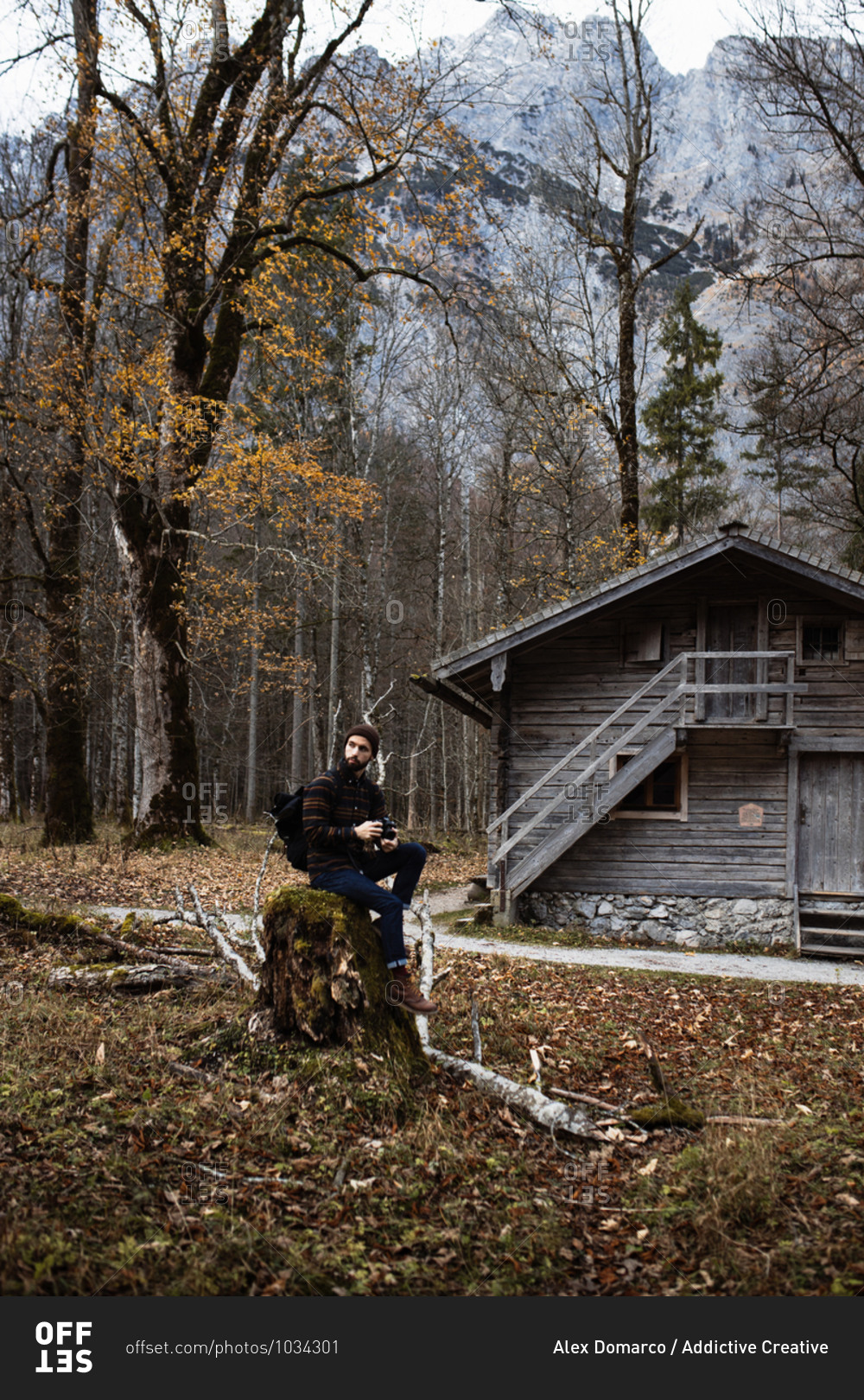 Side view of man in warm outerwear with backpack sitting on stone near old wooden hut located in leafless forest surrounded by mountains in overcast autumn day