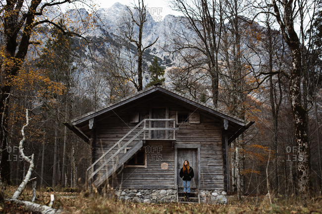 Low angle of young female tourist in warm jacket looking away while standing against shabby lumber exterior of aged building in cold day in countryside