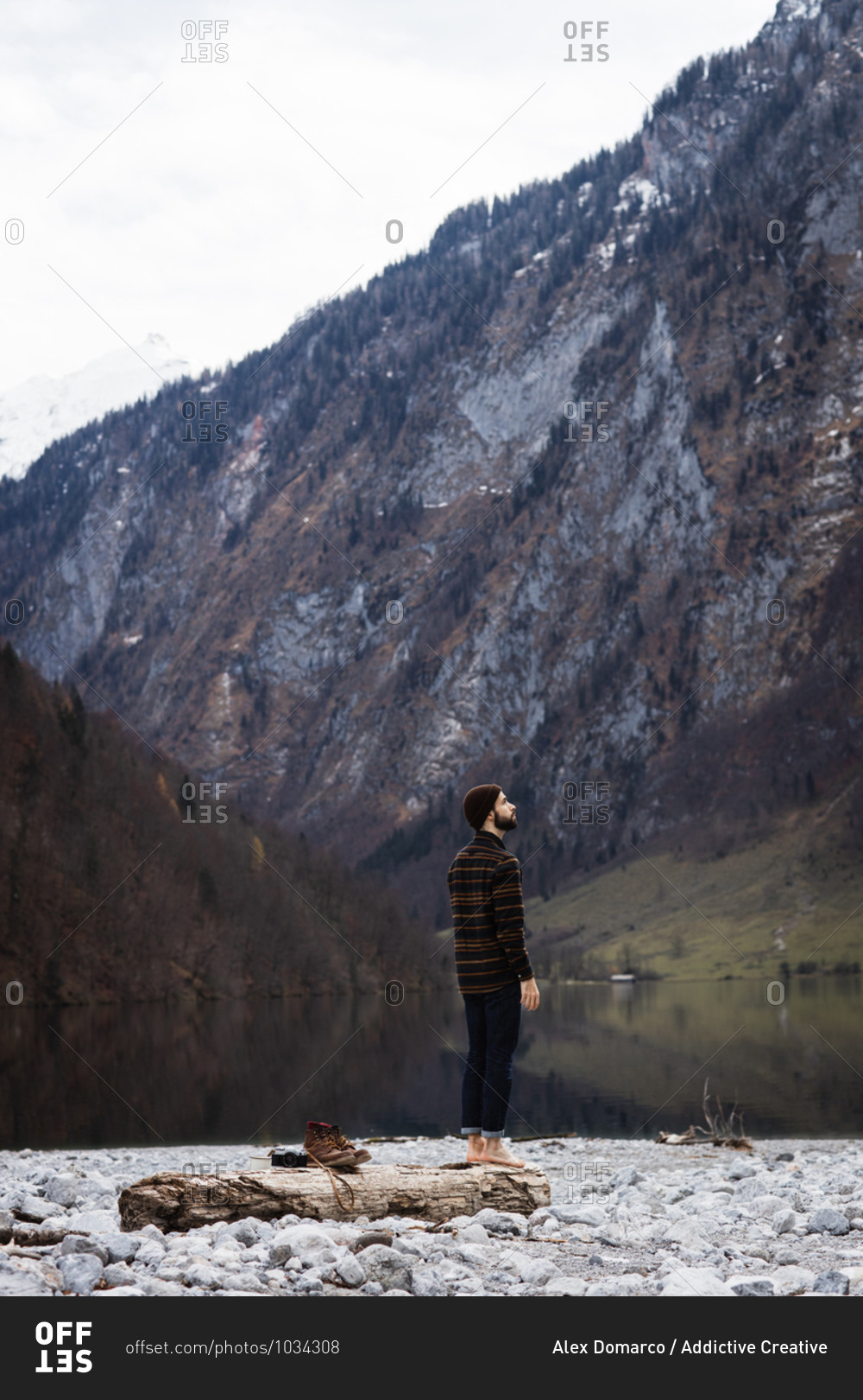 Side view of male tourist enjoying amazing landscape of calm lake and majestic mountains with forested slopes and snowy peaks in cloudy autumn day
