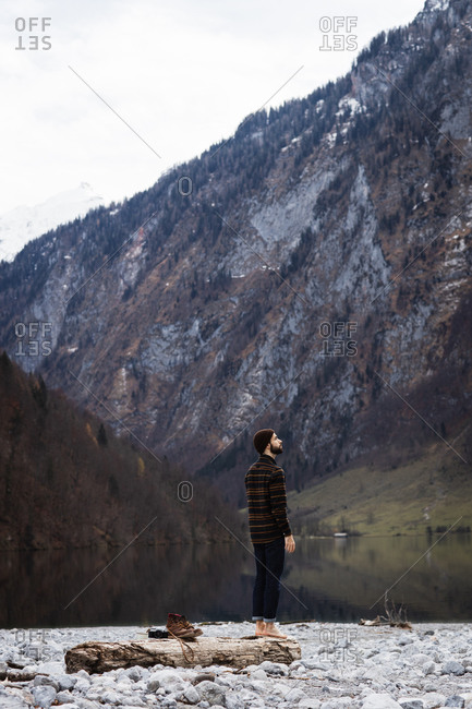 Side view of male tourist enjoying amazing landscape of calm lake and majestic mountains with forested slopes and snowy peaks in cloudy autumn day