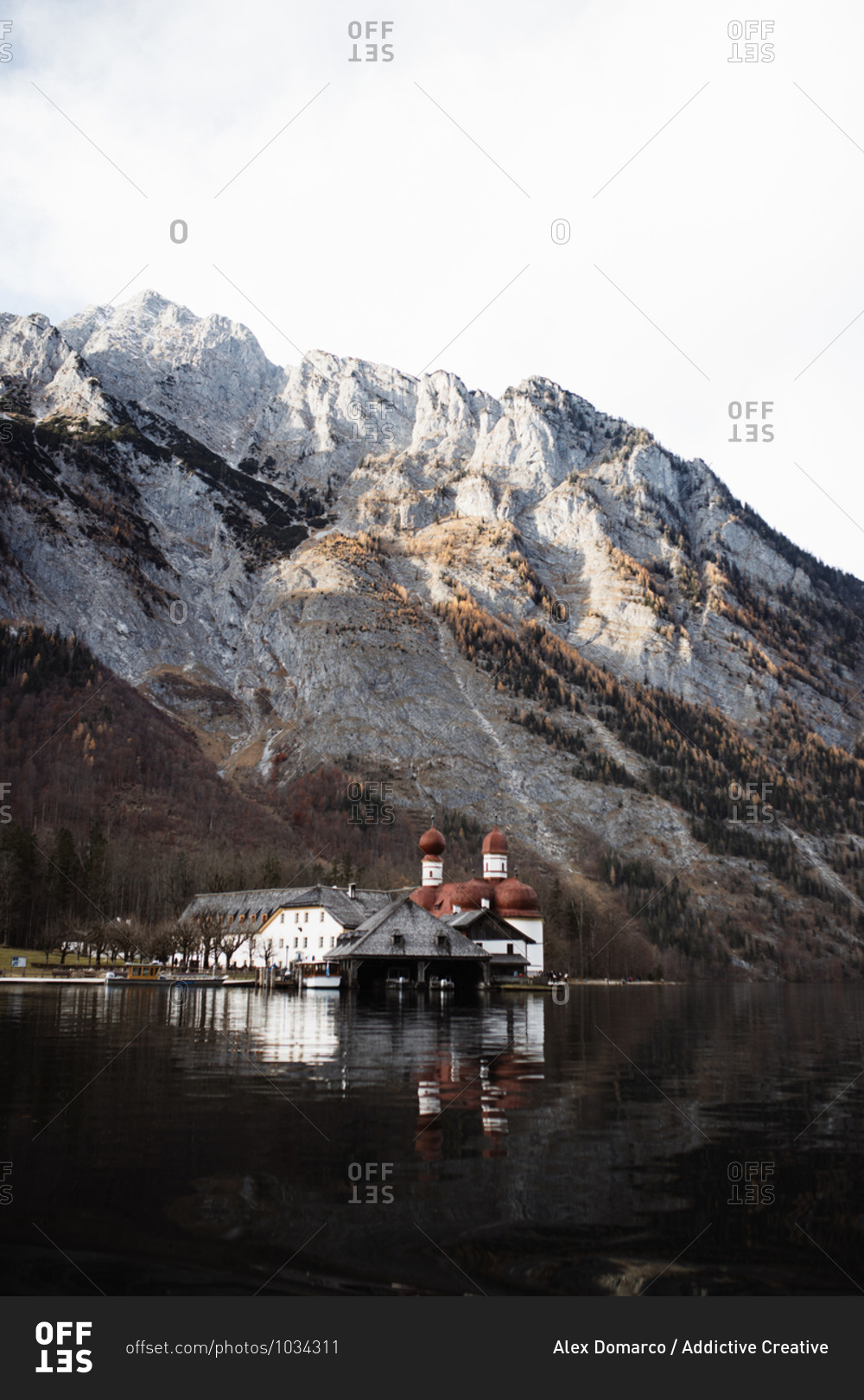 Picturesque landscape with small village houses and church\
located on shore of calm lake against rough rocky slope of mountain\
covered with snow in sunny autumn weather stock photo -\
OFFSET