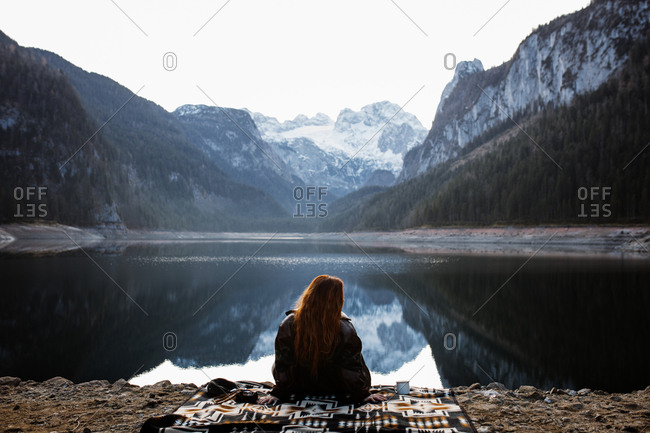 Back view of unrecognizable female traveler sitting on blanket near calm lake surrounded by majestic rocky mountains covered with forest and snow and enjoying silence in autumn morning