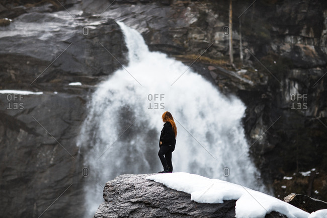 Side view of traveling woman standing on rock and observing amazing view of powerful waterfall in highlands in winter
