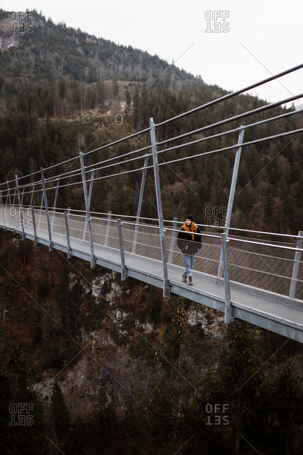 Female tourist in warm clothes walking along metal suspension bridge in highlands and enjoying vacation in autumn