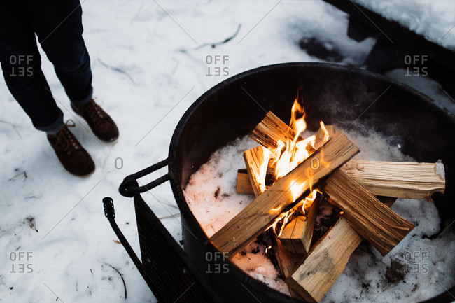 High angle of crop unrecognizable traveler standing on snow and warming up near camping fireplace with burning firewood in winter day