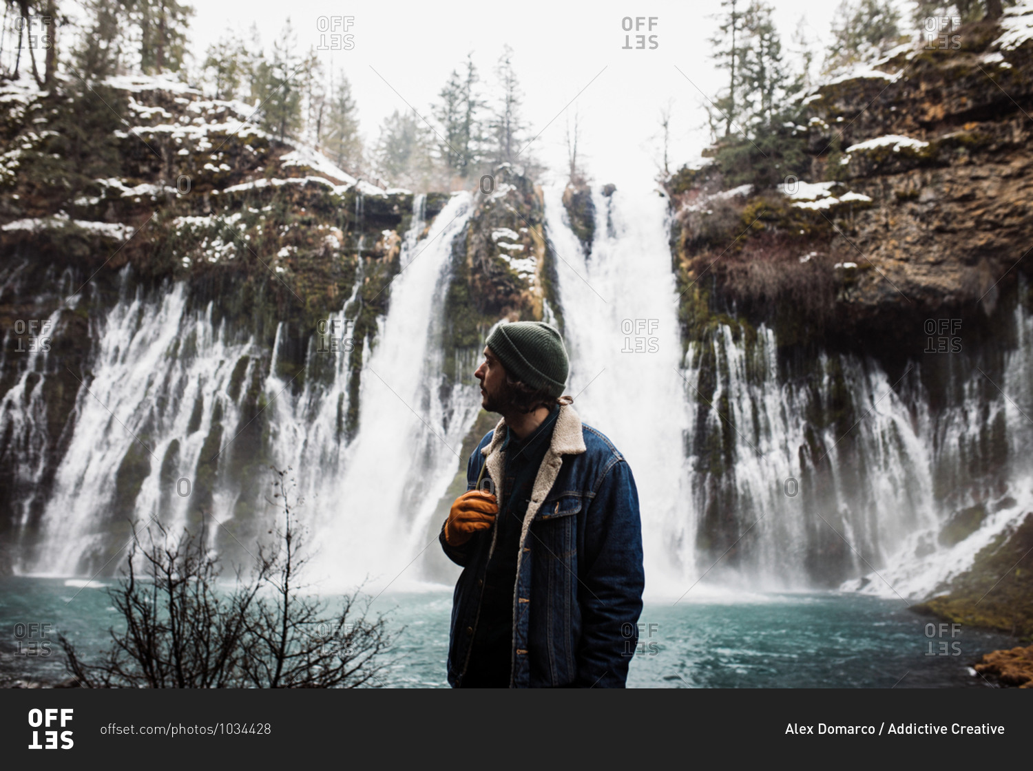 Man standing on picturesque scenery of powerful waterfall with pool flowing among snowy forest in mountainous terrain in winter day in USA looking away