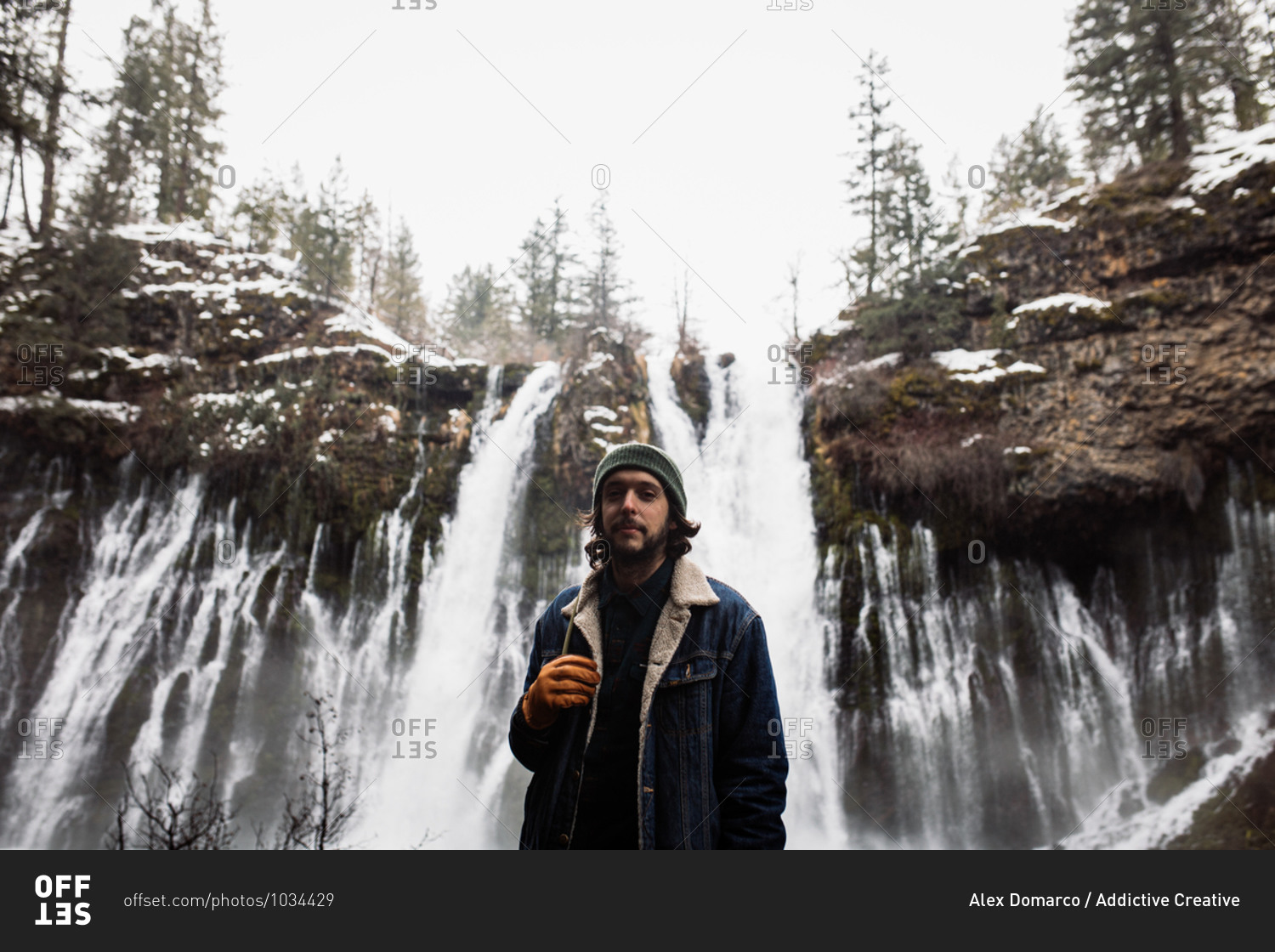 Man standing on picturesque scenery of powerful waterfall with pool flowing among snowy forest in mountainous terrain in winter day in USA looking at camera