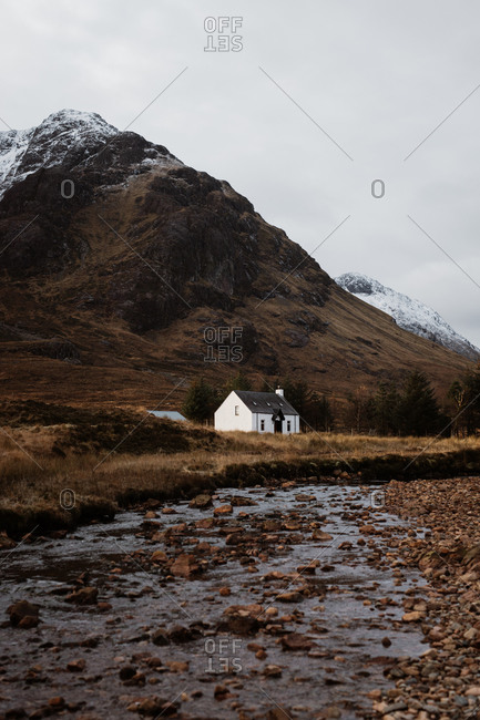 Picturesque view of snowy mountain range and lonely residential house near calm creek under gray sky in Scottish Highlands