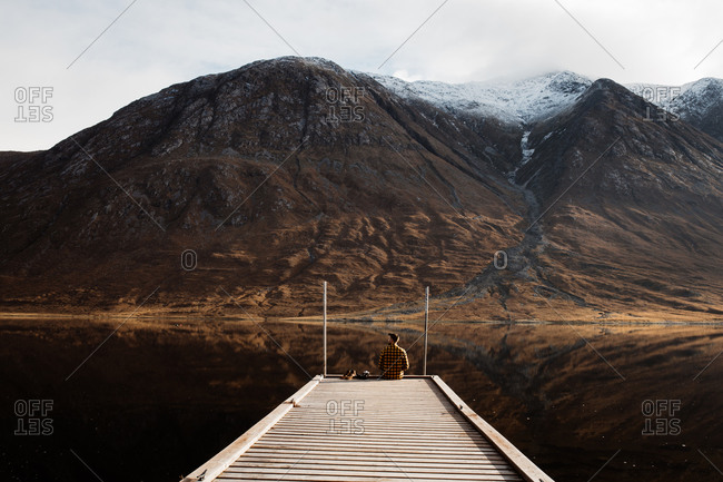 Distant view of lonely tourist sitting on wooden pier and admiring peaceful scenery of mountains and lake on overcast day in Scottish Highlands