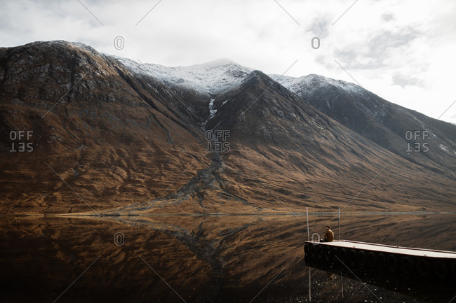 Distant view of lonely tourist sitting on wooden pier and admiring peaceful scenery of mountains and lake on overcast day in Scottish Highlands