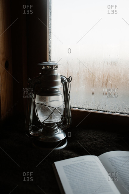 Cozy atmosphere of room with retro oil lantern and opened book placed on windowsill with misted window in house