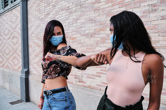 Multiethnic female friends wearing medical masks bumping elbows while greeting each other during coronavirus epidemic