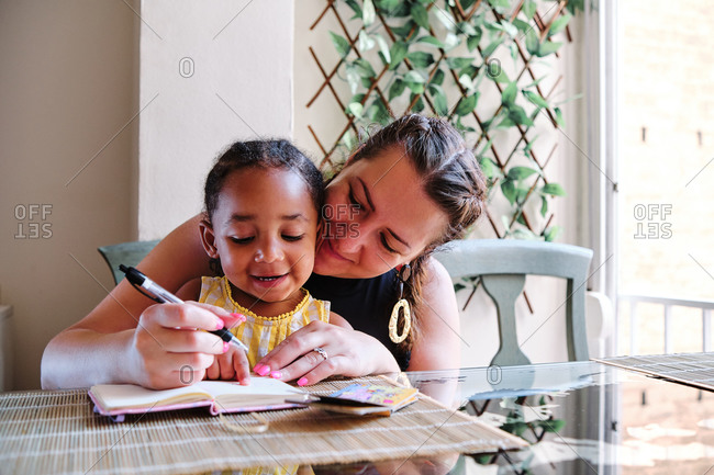 Multiethnic mother and little daughter sitting at table at home and writing in notebook while studying at home