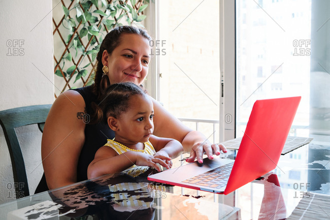 Multiethnic mother and child sitting together at table and watching interesting cartoon on netbook while relaxing at home