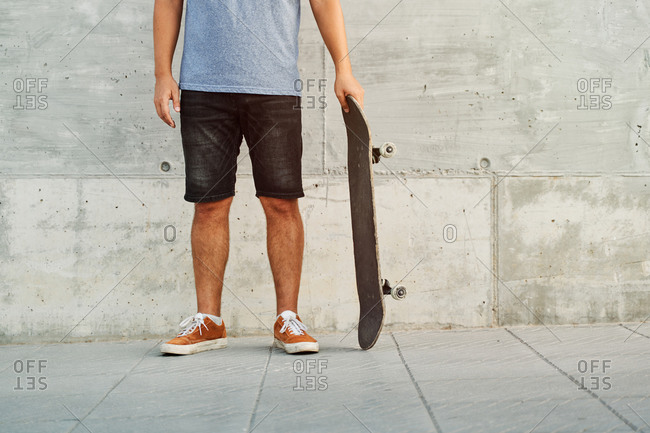 Unrecognizable crop male in street style outfit standing with skateboard near urban building in city