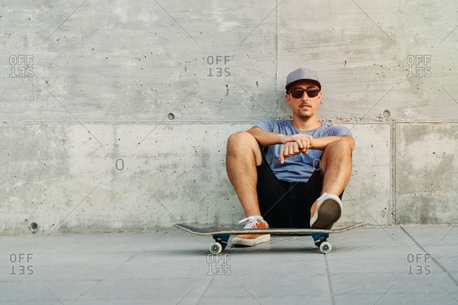 Confident male skater in stylish outfit sitting near urban building with skateboard and looking at camera