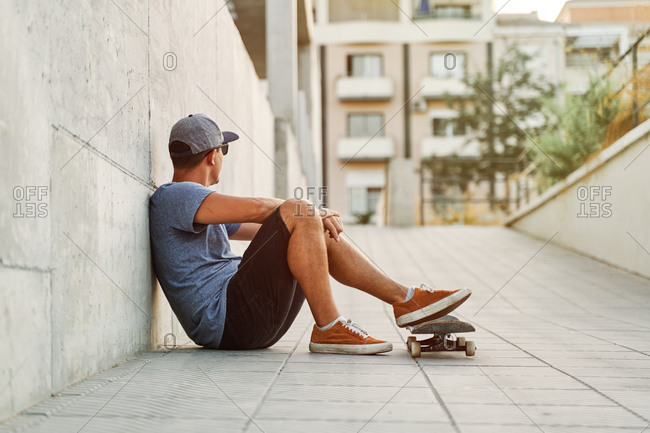 Confident male skater in stylish outfit sitting near urban building with skateboard and looking away