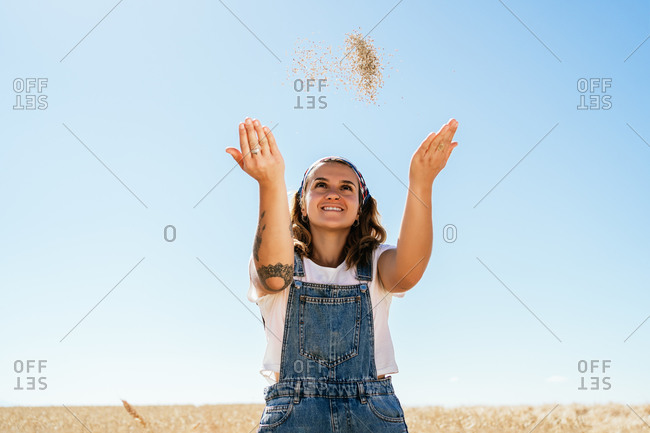 Low angle of delighted female tossing up wheat grain while standing in agricultural field in sunny day in village