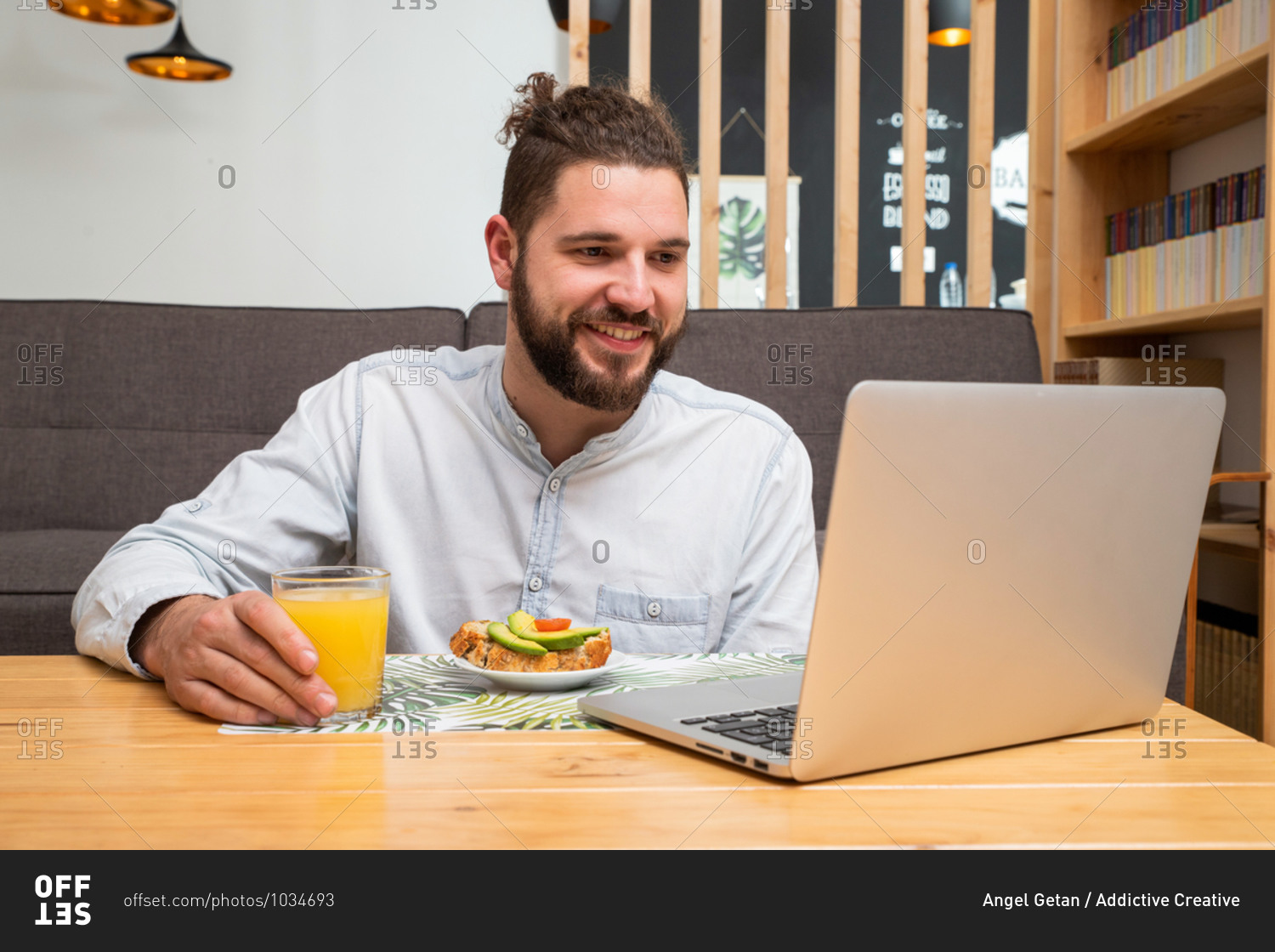 Cheerful male entrepreneur sitting at table with glass of juice and vegetable toast while reading documents on laptop and working remotely