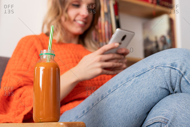 Low angle of bottle of detox smoothie placed on table on background of female sitting on sofa and browsing cellphone