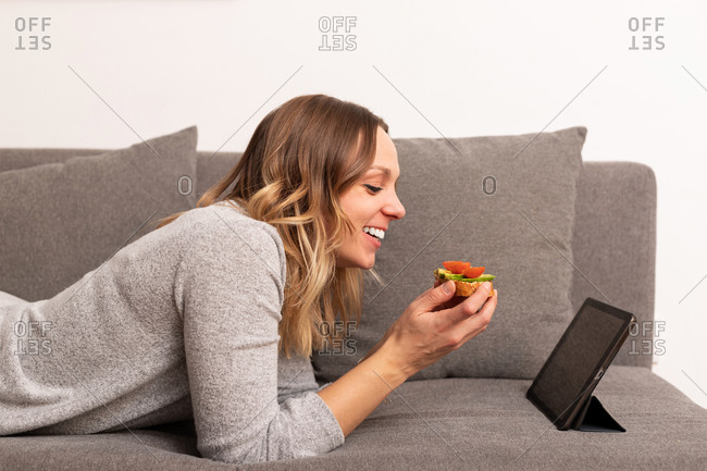 Side view of cheerful female eating healthy toast with vegetables while watching movie on tablet at home and lying on sofa