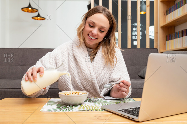 Positive female in bathrobe pouring milk in bowl with cereal while sitting at table with laptop and having nutritious breakfast