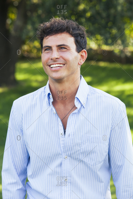Happy handsome young male in unbuttoned white shirt looking away confidently while standing against green plants in summer garden