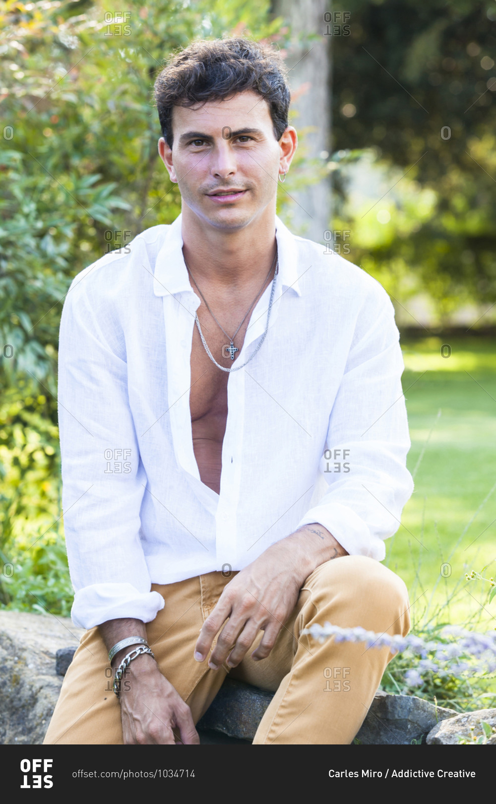 Happy young male in unbuttoned white shirt and colorful jeans with accessories sitting on stone border in green summer garden