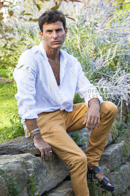 Full length of happy young male in unbuttoned white shirt and colorful jeans with accessories sitting on stone border in green summer garden