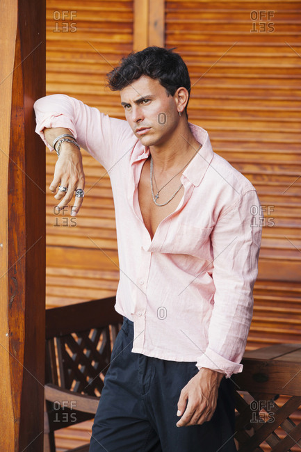 Confident young casually dressed male in unbuttoned shirt and pants leaning on pillar and looking away pensively while standing on rustic terrace with wooden furniture