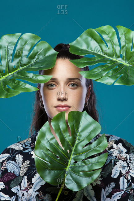 Young female model hiding face behind green monstera leaves looking at camera in studio with turquoise background