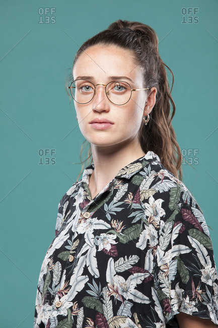 Side view of teen female model in eyeglasses dressed in casual shirt with tropical print and jeans standing in blue studio and looking at camera