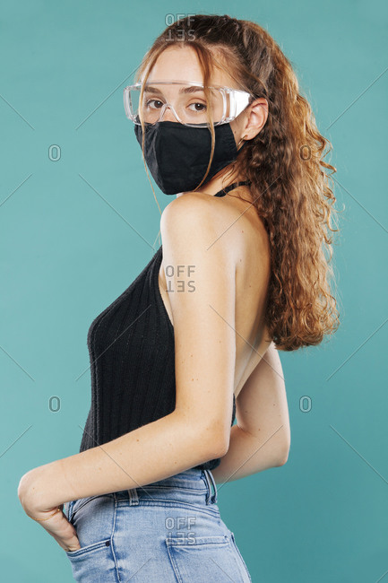 Side view of female wearing medical mask and plastic safety goggles standing on blue background in studio and looking at camera