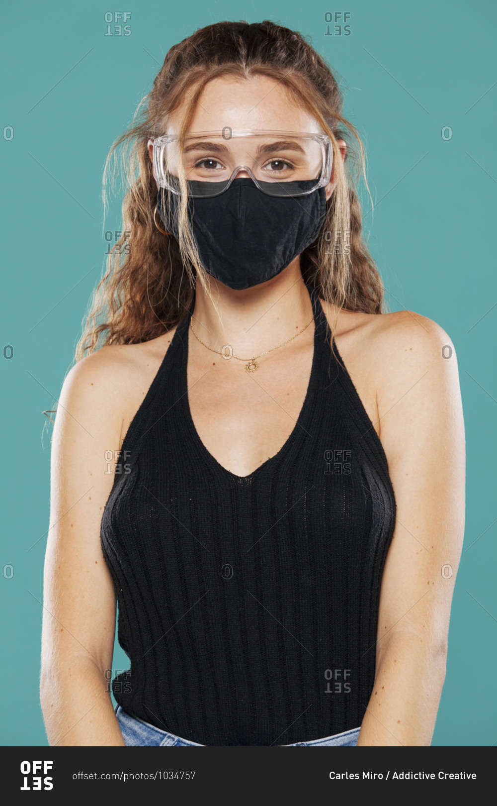 Female wearing medical mask and plastic safety goggles standing on blue background in studio and looking at camera